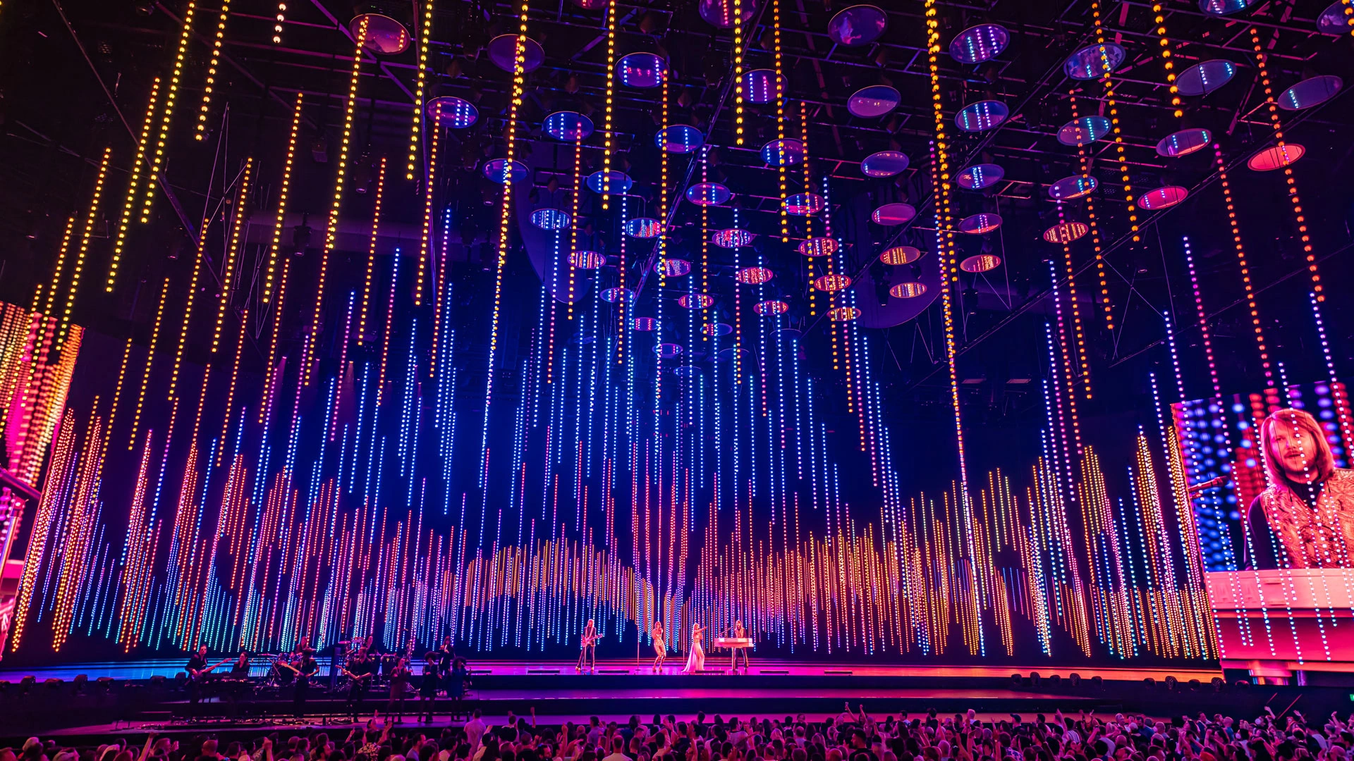 Theatrical performance with overhead suspended lighting, colourful visual effects and an audience in the foreground